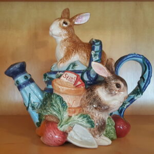 A hand-painted Fitz & Floyd teapot in the shape of a blue watering can surrounded by three bunnies in a garden of vegetables. The brown and white rabbits appear to be nibbling or playing near a seeds packet, all finished with glossy glaze and detailed fur texture.