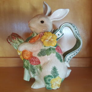 Front view of a 1996 Fitz and Floyd “Herb Garden” Bunny Teapot featuring a hand-painted ceramic rabbit holding a carrot and artichoke, surrounded by pansies and greenery. The handle is shaped like a green ribbon with “Parsley, Thyme, Basil” written on it. The teapot has a glossy finish and vibrant spring colors, set against a plain background.